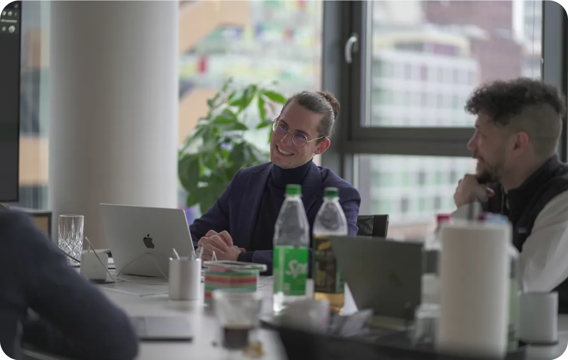 A focused team meeting with laptops, beverages, and note-taking, illuminated by natural light in an office setting.