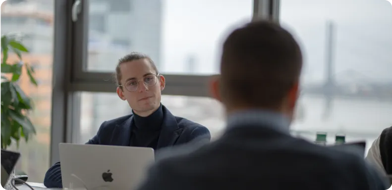 A focused individual with glasses in a business-casual outfit sits in front of a MacBook, facing another person across the desk in an office with a large window overlooking a river and cityscape.