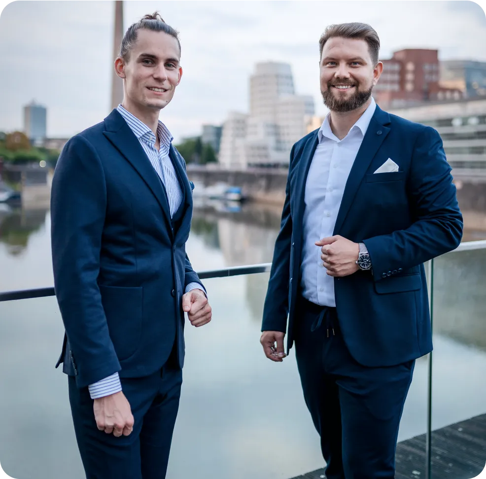 wo professional men in business attire smiling and standing on a balcony with a city riverfront backdrop