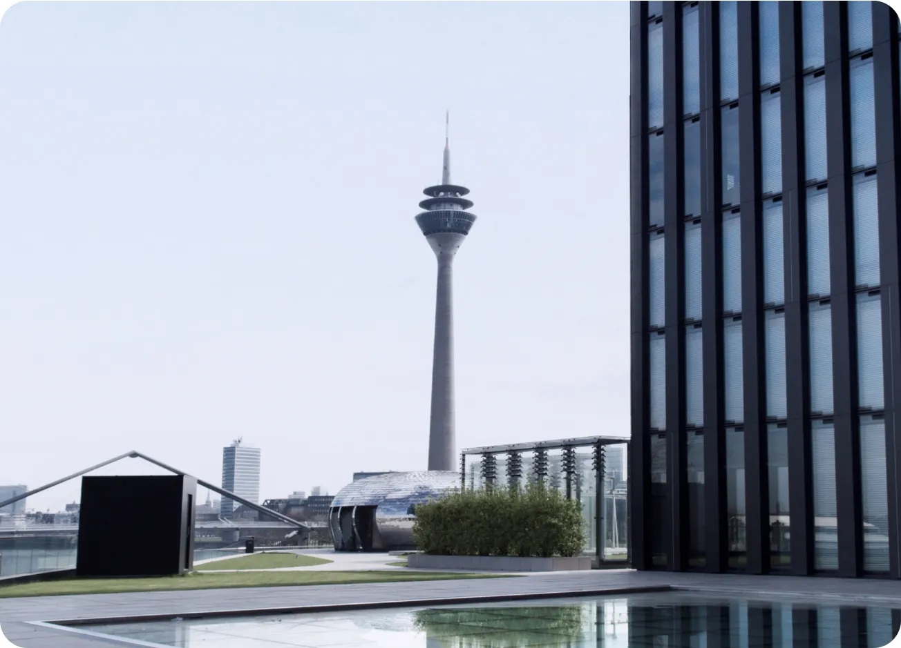 A modern cityscape with a distinctive tower in the background, viewed from a terrace with a reflective pool and an architectural glass building on the right.